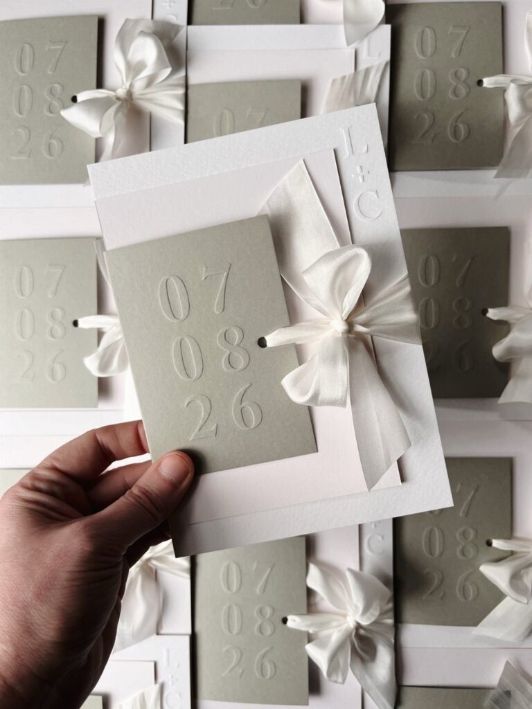 Hand holding a white wedding invitation with a grey embossed date card and ribbon.