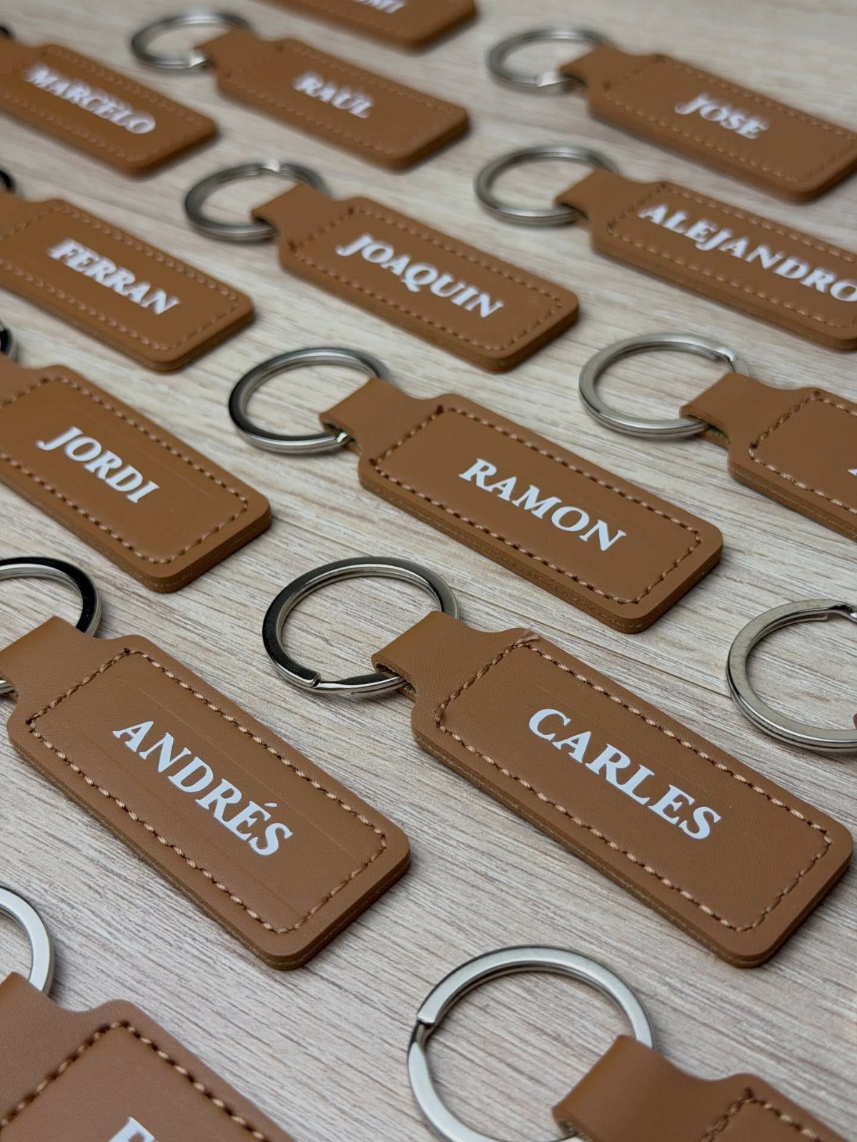 Brown leather keychains with silver rings and white names on a wooden surface.