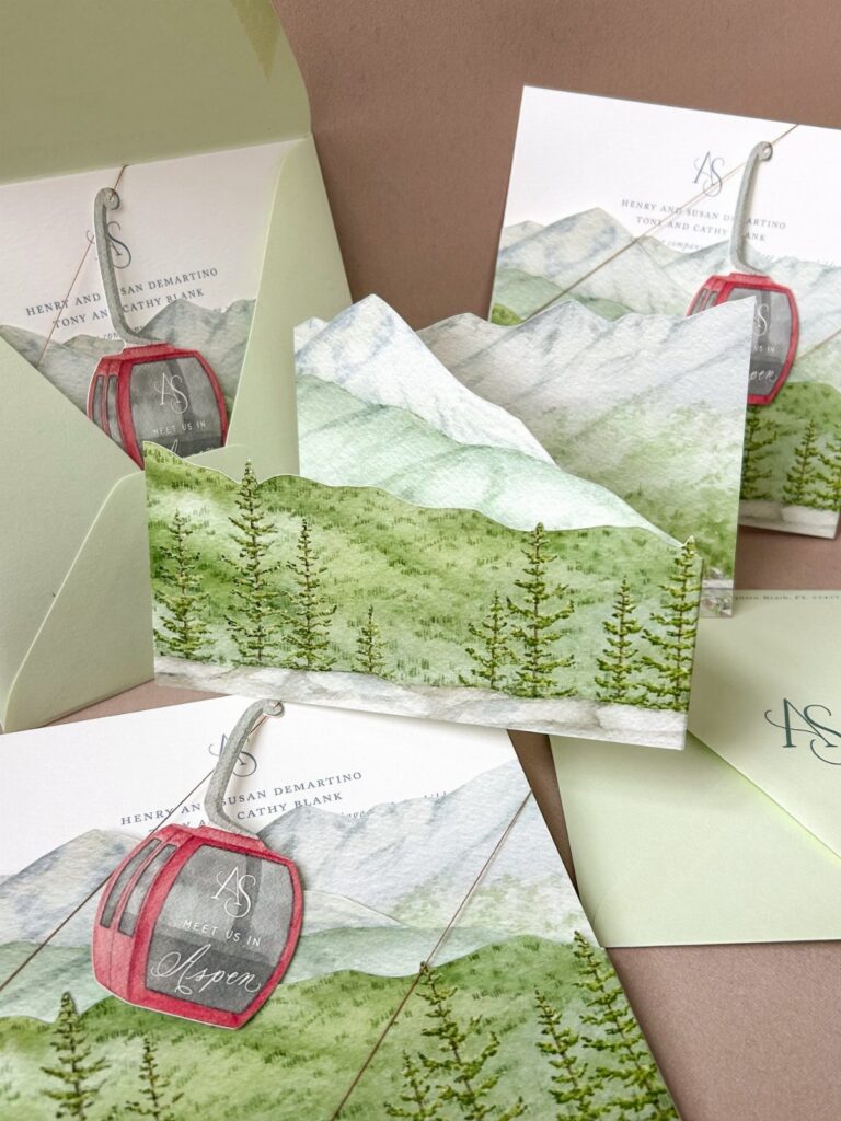 Wedding invitations with watercolor mountains, pine trees, and red gondolas with green envelopes.
