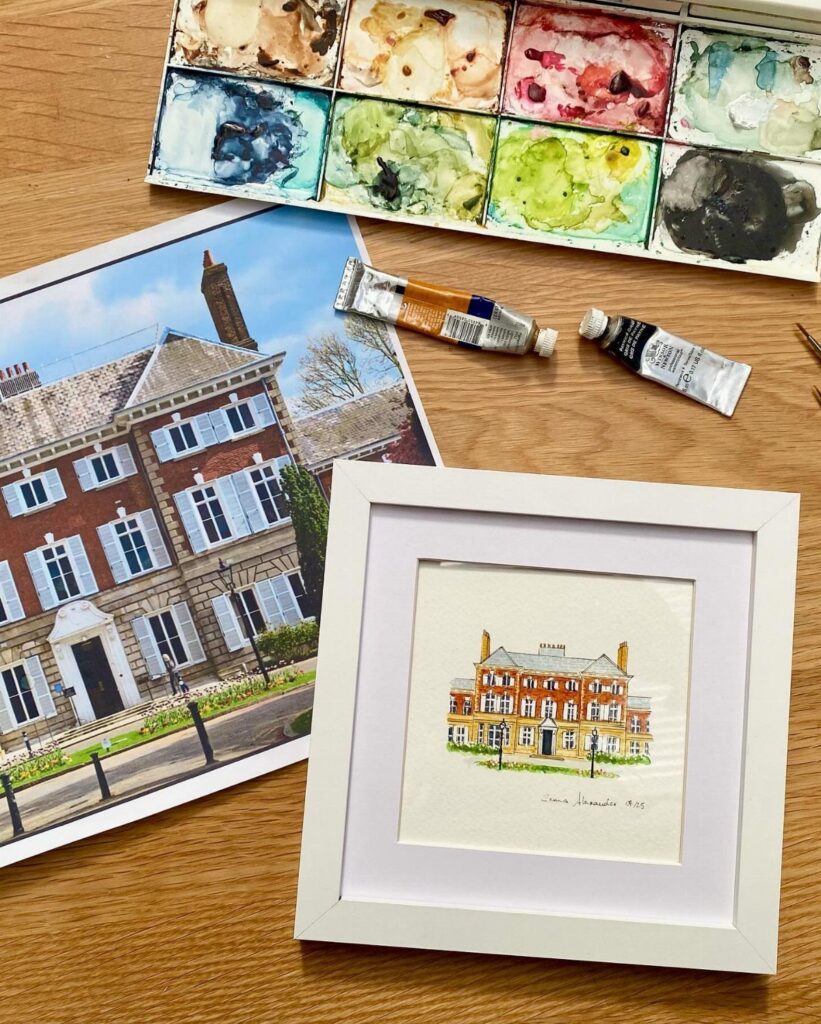 Watercolor painting of a brick house in a white frame with art supplies.