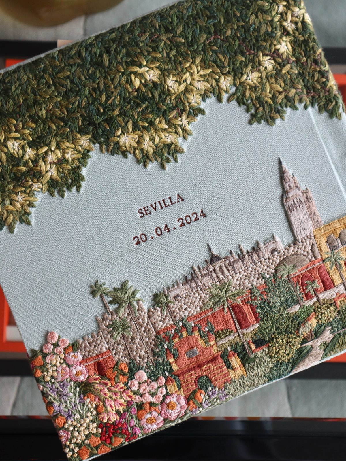 Embroidered fabric cover showing a Sevilla landscape with date and city name stitched.