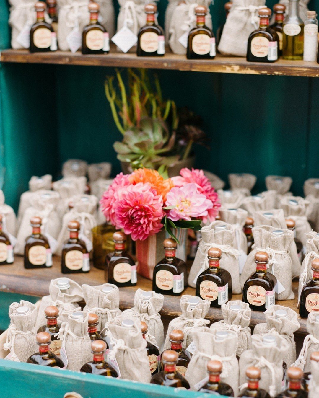 Mini tequila bottles in burlap sacks arranged on teal shelves with pink flowers