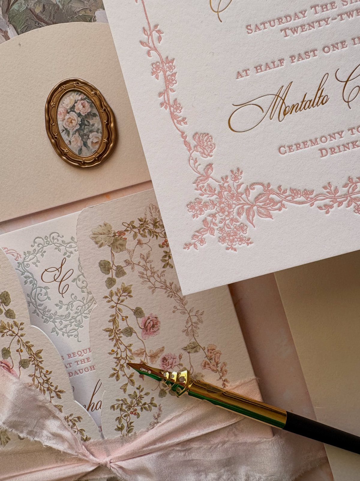Elegant wedding invitation stationery with pink floral details, a gold cameo, and fountain pen.