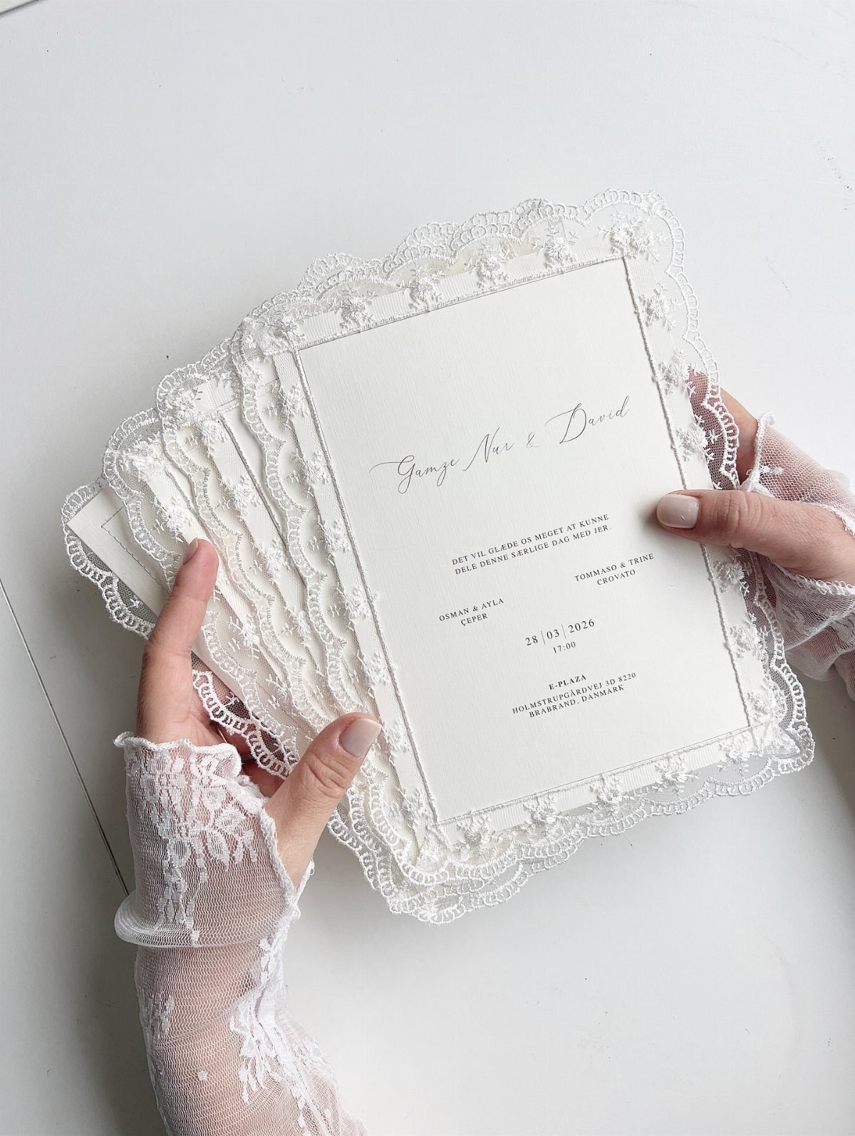 White lace wedding invitations held by hands wearing matching white lace sleeves.