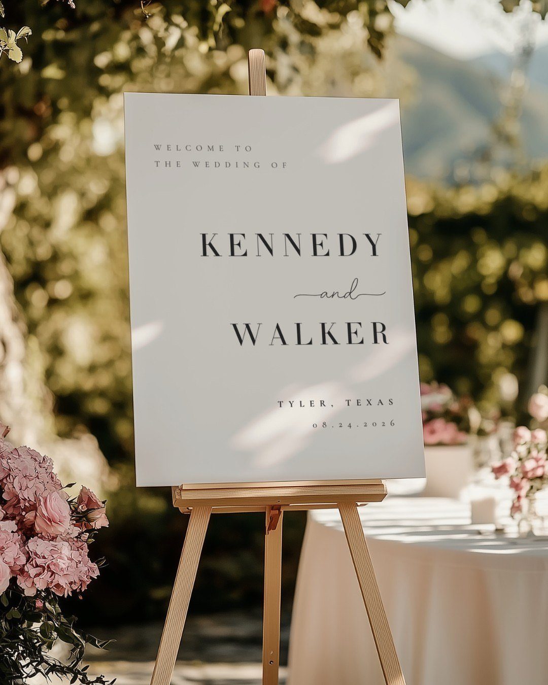 White wedding welcome sign on a wooden easel surrounded by greenery and pink flowers.