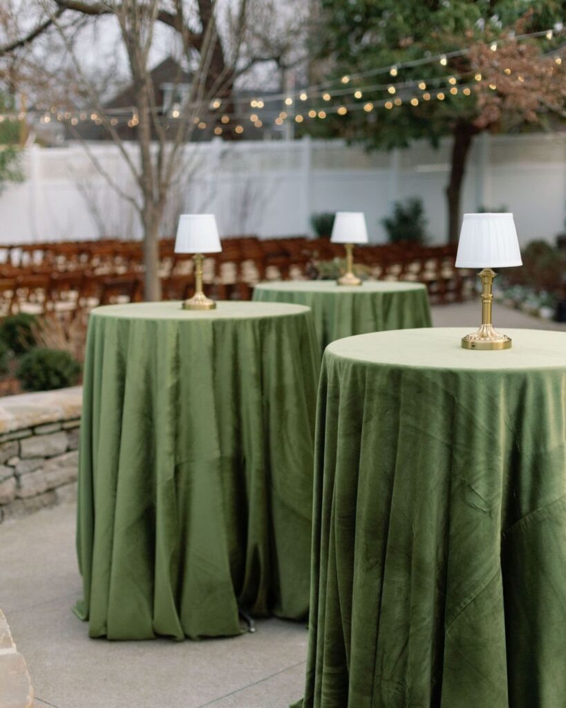 Olive green velvet cocktail tables with gold lamps in an outdoor wedding setting.